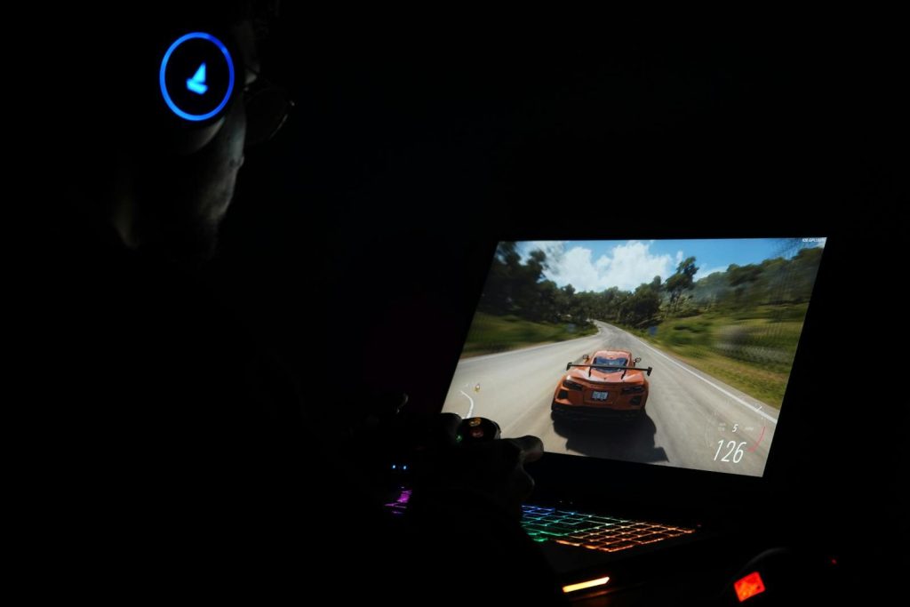 A gamer using a laptop and headphones to play a car racing game in a dark setting.