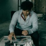 A man in a button-up shirt thinks about his chess move at a wooden table in a dimly lit room.