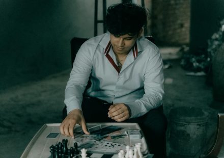 A man in a button-up shirt thinks about his chess move at a wooden table in a dimly lit room.