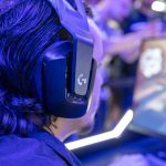 Focused gamer with headphones playing a video game on a PC under blue lighting in a crowded room.