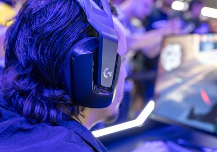 Focused gamer with headphones playing a video game on a PC under blue lighting in a crowded room.