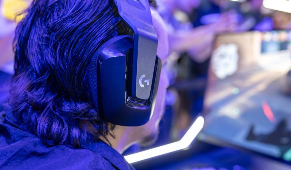Focused gamer with headphones playing a video game on a PC under blue lighting in a crowded room.