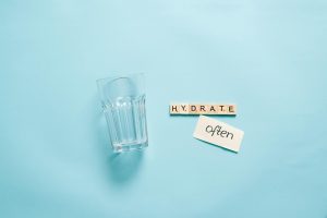A minimalist composition of a glass with Scrabble tiles spelling 'HYDRATE' on a blue background.
