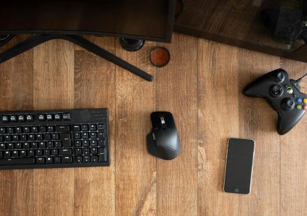 Top view of black gamepad with mouse and keyboard on wooden desk with monitor in apartment in daytime