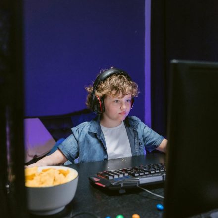 A child wearing headphones intensely playing video games in a dimly lit room with snacks nearby.