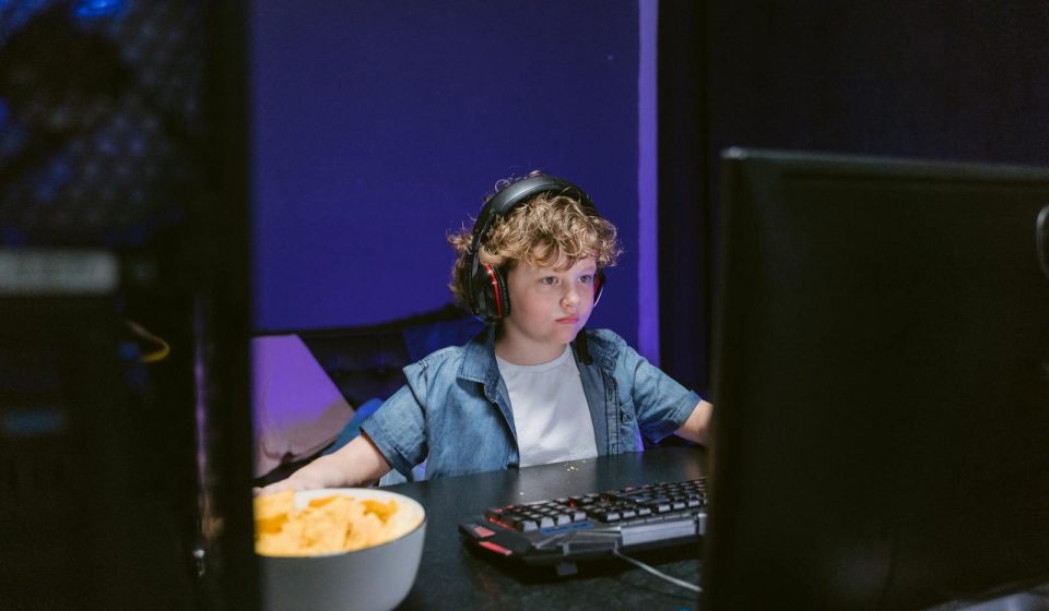 A child wearing headphones intensely playing video games in a dimly lit room with snacks nearby.