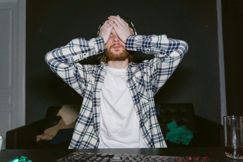 Focused male gamer in plaid shirt, showing stress with hands on face, indoors.