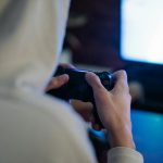 Back view of crop anonymous man with console controller playing video game against screen at home