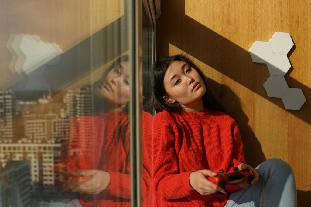 A young woman in a red sweater plays video games by a window in an urban apartment.