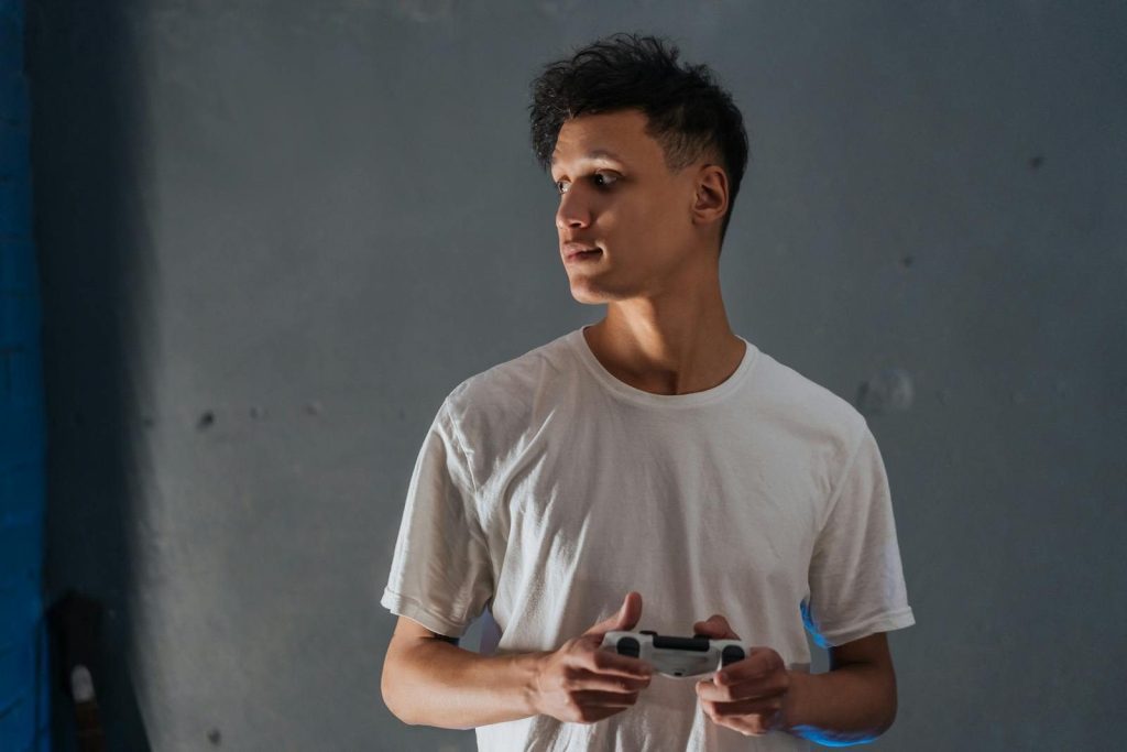 A young man wearing a white t-shirt focuses intently while holding a game controller indoors.