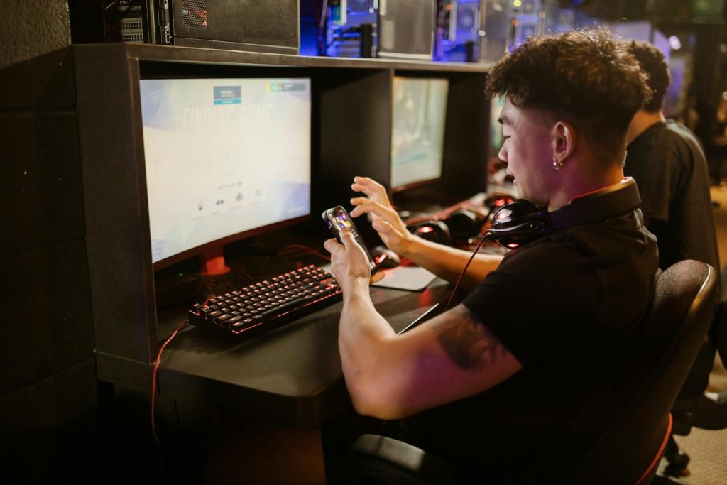A young man is gaming using a smartphone and a PC setup with a mechanical keyboard indoors.