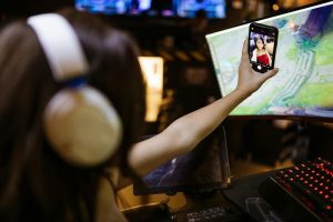 A woman wearing headphones takes a selfie with her smartphone while engaged in gaming indoors.