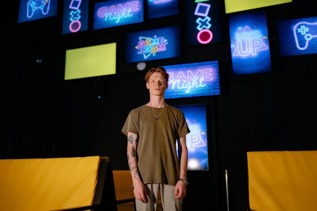 A young man stands in a game room, surrounded by colorful neon signs.