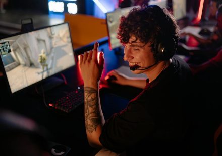 A young gamer in a black shirt enjoys an intense online game on a computer, smiling confidently.