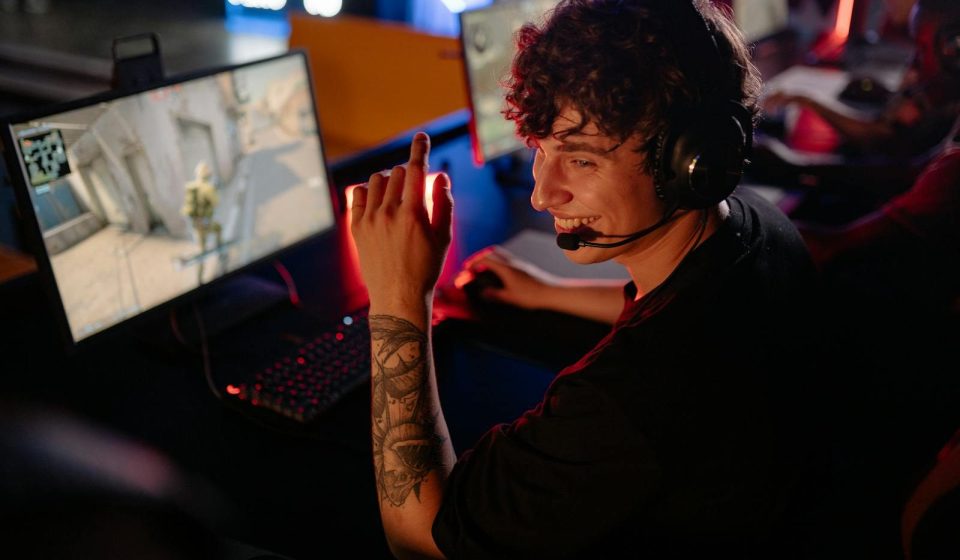 A young gamer in a black shirt enjoys an intense online game on a computer, smiling confidently.
